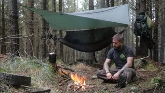un homme assis près d'un feu bivouac dans la foret. Derrière lui, un hamac avec sa moustiquaire et son tarp