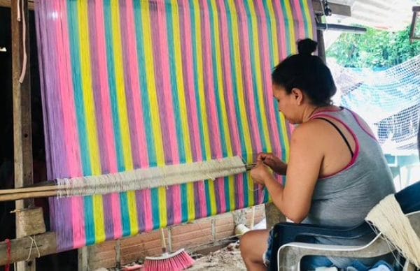 photo d'une artisane Colombienne assise sur une chaise, qui tisse à la main, un hamac multicolore, sur un métier à tisser verticale. 